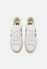 Λευκά δερμάτινα αθλητικά παπούτσια με μπλε λεπτομέρειες, στρογγυλή μύτη και επίπεδα κορδόνια. Ανάγλυφη επιφάνεια, ορατά ραφή και καφέ καουτσούκ σόλα.