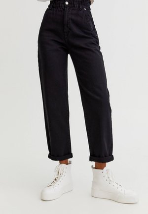 Onderlichaam gekleed in zwart, hooggetailleerde jeans met opgerolde boorden en witte, hoge platform sneakers, tegen een effen achtergrond.