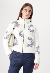Weiße Kunstfelljacke mit floraler Stickerei in Lila und Blau, mit hohem Kragen und Frontreißverschluss, kombiniert mit navyfarbenen Hosen.