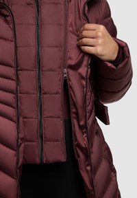 Vijolična puhovka z vodoravnim prešitjem, sprednjo zadrgo in nastavljivimi manšetami. Gladek, sijoč material z mehko teksturo.