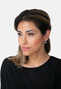 Porträt einer Frau mit schulterlangen braunen Haaren und Strähnchen, die ein schwarzes Shirt, Ohrringe in Form von Ringen und dezentes Make-up mit einem natürlichen Look trägt.