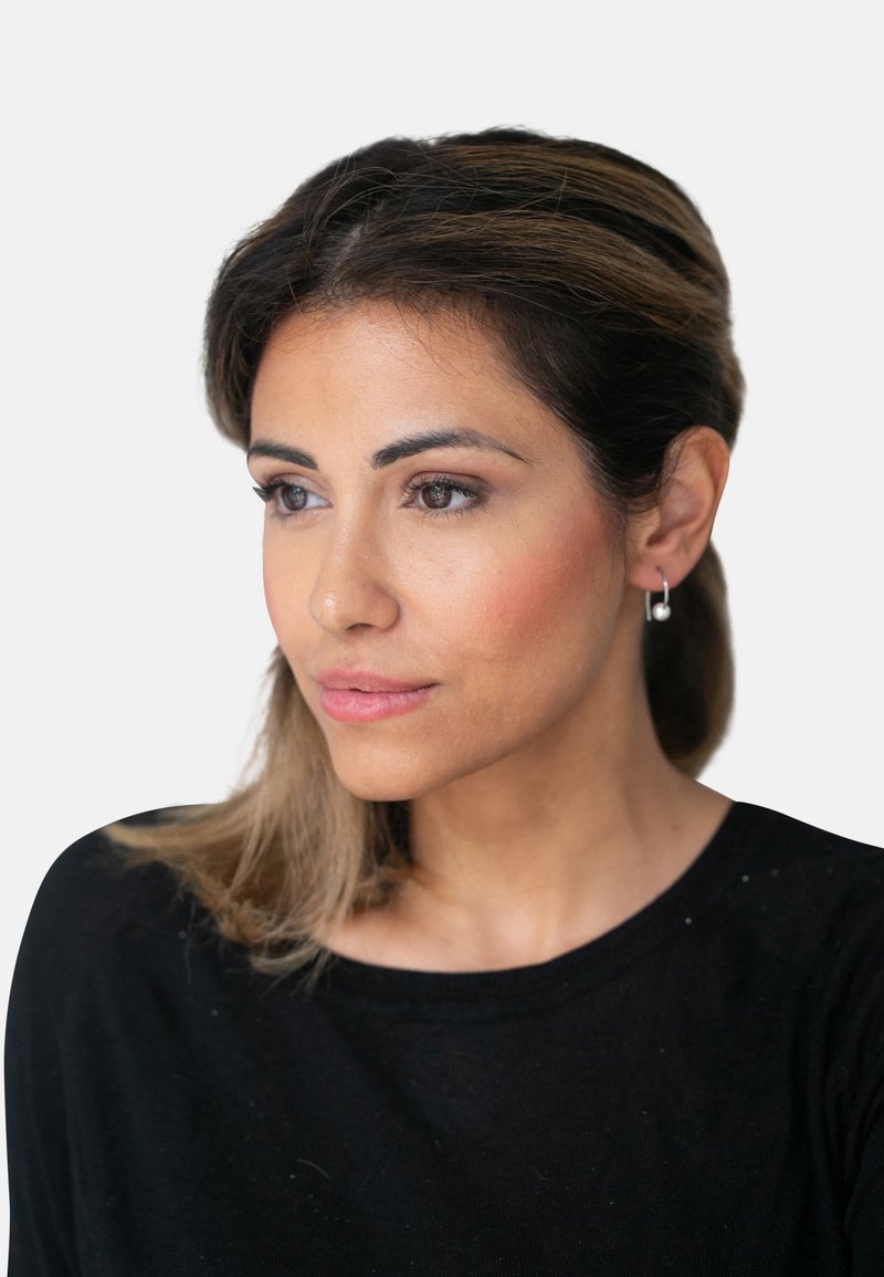 Porträt einer Frau mit schulterlangen braunen Haaren und Strähnchen, die ein schwarzes Shirt, Ohrringe in Form von Ringen und dezentes Make-up mit einem natürlichen Look trägt.