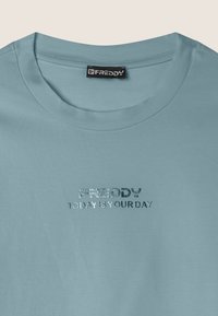 T-shirt in cotone azzurro chiaro con scollo rotondo e testo in argento metallico: "FREDDY OGGI È IL TUO GIORNO" sul davanti.