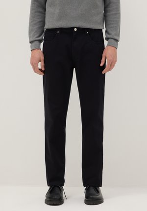 Hombre con pantalones rectos negros, zapatos negros y un suéter gris de pie contra un fondo claro y liso.