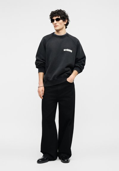 Άνδρας που φοράει μαύρο φαρδύ φούτερ με λευκό λογότυπο, μαύρο παντελόνι φαρδιάς γραμμής, μαύρα παπούτσια, γυαλιά ηλίου, στέκεται με το ένα χέρι στην τσέπη.