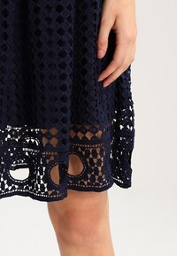 Námořnický modrý šaty z jemné krajky s háčkováním, s geometrickým vzorem a kruhovými výřezy po dolním okraji. Ruka viditelně drží látku.