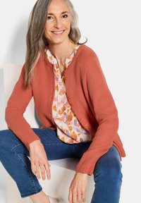 Cardigan en tricot corail avec des poignets côtelés, associé à une chemise à motifs floraux. Le modèle porte un jean bleu et est assis contre un fond clair.