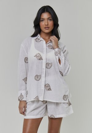Hvid linnedskjorte med brun skalkbroderi og knapper, kombineret med matchende shorts med samme skalmønster. Letvægtsstof.