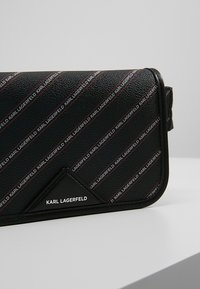 Borsa a tracolla nera con superficie testurizzata, caratterizzata da strisce con logo a motivo in bianco e rosso, e un accento triangolare con il nome del marchio.