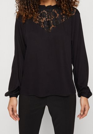 Femme portant un chemisier noir à manches longues avec un détail en dentelle florale sur le haut de la poitrine et un pantalon noir ajusté, debout devant un fond uni.