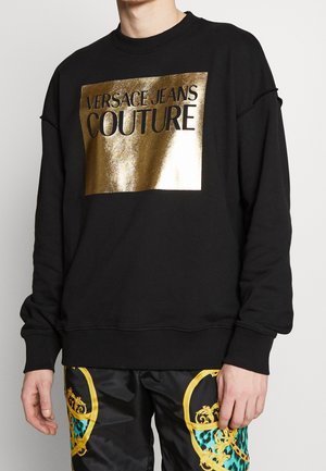 Persona che indossa una felpa nera con la scritta dorata "Versace Jeans Couture" e pantaloni neri con motivi barocchi ornati in oro e teal.