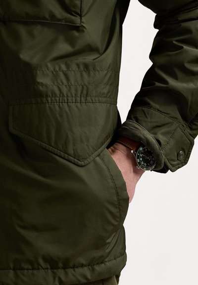 Dunkelgrüne gepolsterte Jacke mit einer Seitentasche und Knopfmanschette, kombiniert mit einer Uhr mit schwarzem Zifferblatt und grünen Akzenten.