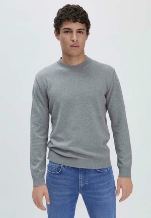 Grauer Strickpullover mit Rundhalsausschnitt und gerippten Bündchen, kombiniert mit blauen Jeans. Der Stoff wirkt glatt und leicht.