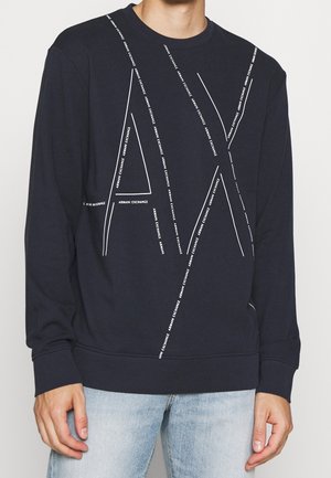 Man wearing navy sweatshirt with large white "AX" lines and repeated "Armani Exchange" text, paired with light blue jeans.