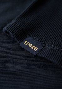 Mørkeblå ribbet sweaterstof med et vævet mærke, der viser "SUPERDRY" i gyldne bogstaver, med en glat tekstur og subtil glans.