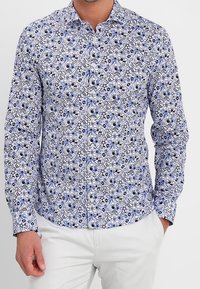 Camicia bianca con motivi floreali e astratti blu, maniche lunghe, colletto button-down e design slim fit. Indossata con pantaloni di colore chiaro.