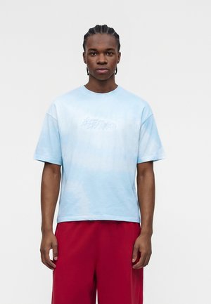 Mladý muž s vrkočmi, oblečený v svetlomodré nadrozmerné tričko a červené voľné nohavice, stojaci pred jednoduchým svetlým pozadím.