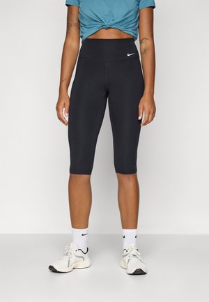 Sorte capri leggings med høj talje, glat tekstur, prydet med et lille hvidt Nike-logo. Båret med en tætsiddende blå top og hvide sneakers.