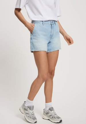 A person stands wearing a white t-shirt and light blue denim shorts, paired with grey sneakers and white socks.