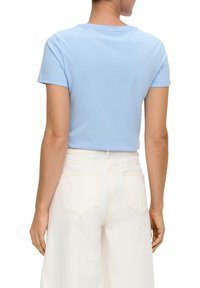 Himmelblaues Baumwoll-T-Shirt mit kurzen Ärmeln und rundem Ausschnitt, von hinten gezeigt. Kombiniert mit hochgeschnittenen weißen Jeans.