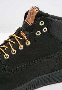 Botín negro de cuero que presenta ojales dorados, cordones amarillos y rojos, y un parche de logo marrón en el cuello. Detalle de la parte superior texturizado visible.