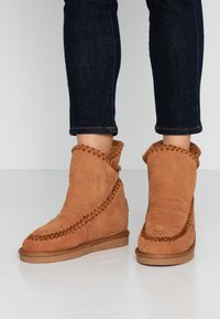 Bottes ankle en suède beige avec un revers replié, des coutures décoratives le long de la couture et une semelle en caoutchouc, associées à un jean en denim foncé.