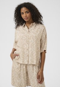 Chemise beige à manches courtes et col, avec un motif abstrait blanc, fabriquée en tissu léger. Comprend des boutons et une coupe décontractée.