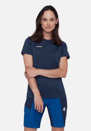 Femme aux longs cheveux bruns portant un t-shirt à manches courtes bleu marine de la marque Mammut et un short bleu Mammut, debout les bras croisés avec une expression neutre.