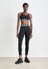 Femme portant un soutien-gorge de sport noir Calvin Klein et un legging taille haute avec des chaussures de sport noires, debout contre un mur clair.