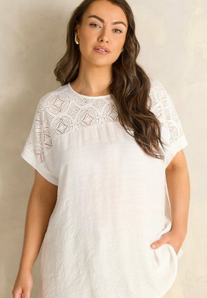 Donna con capelli castani lunghi che indossa una camicetta bianca a maniche corte con inserto in pizzo, posa con una mano in tasca davanti a uno sfondo beige.