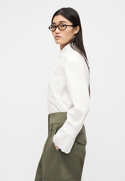 Chemise blanche à manches longues avec une poche et des détails en broderie anglaise sur les poignets, portée avec un pantalon taille haute vert olive. Lunettes marron.