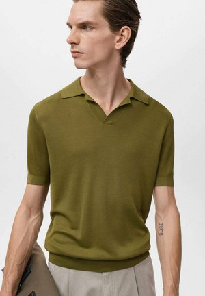 Olivgrünes kurzärmliges Poloshirt mit strukturiertem Strick, V-Ausschnitt und gerippten Bündchen. Getragen mit hellen Hosen und einer gefalteten Mappe in der Hand.