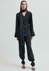 Czarna bluzka z długim rękawem, z dopasowanym szalikiem. Połączona z luźnymi czarnymi spodniami z węzłowymi mankietami.