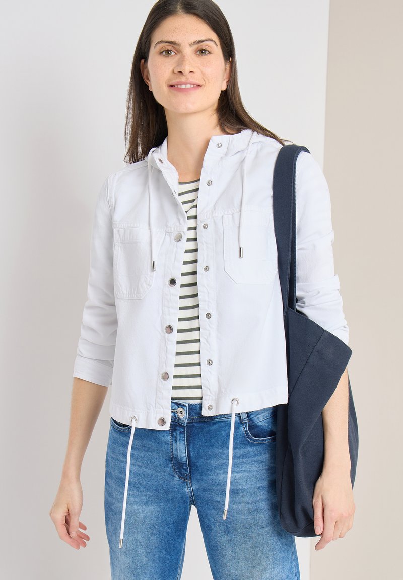 Jeune femme portant une veste blanche boutonnée sur une chemise rayée et un jean bleu, tenant un sac fourre-tout foncé sur son épaule.