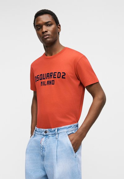 Άνδρας που φοράει πορτοκαλί T-shirt Dsquared2 Milano και ανοιχτό μπλε τζιν, στέκεται με τα χέρια στις τσέπες μπροστά σε απλό φόντο.