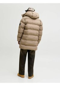 Abrigo acolchado beige con capucha, con secciones acolchadas, una insignia en la manga y un ajuste relajado, combinado con pantalones oscuros y botas color miel.