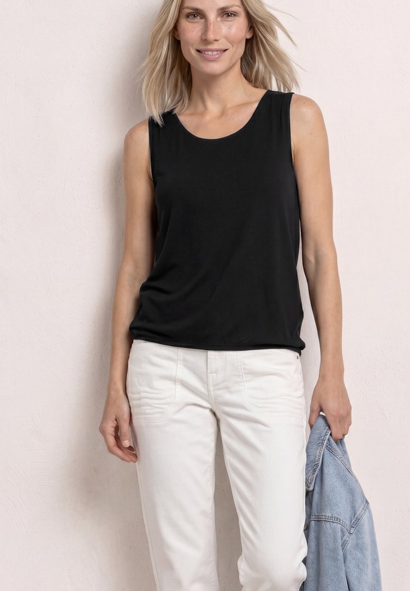 Femme blonde souriante, portant un haut noir sans manches et un pantalon blanc, tenant une veste en jean bleu clair dans la main droite.