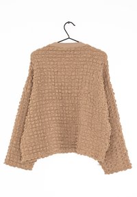 Cardigan beige texturé avec une coupe ample, présentant un motif en nid d'abeille et des manches larges. Suspendu à un crochet noir.