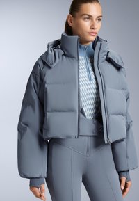 Svijetlosiva skraćena puffer jakna s visokim ovratnikom, velikim rukavima i gumbima, složena preko uzorčane plave majice i odgovarajućih tajica.
