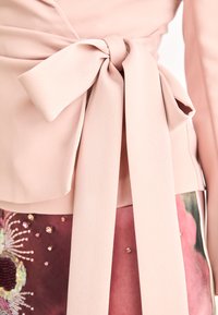 Blush-farget topp med en stor sløyfedetalj i midjen, laget av glatt stoff, kombinert med et mønstret skjørt dekorert med pynt.