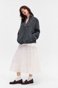 Casaco cinza tricotado com fecho éclair, tecido texturizado, dois bolsos laterais. Vestido midi branco em camadas com rendas, combinado com mocassins burgundy e meias.