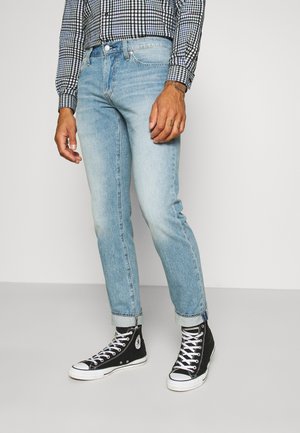 Volwassene draagt lichtblauwe opgerolde spijkerbroek, zwarte hoge sneakers en een zwart-wit geruit shirt met lange mouwen, staand op een witte vloer.