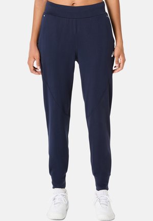 Personne portant un pantalon de sport bleu marine et des chaussures de sport blanches, debout, les mains détendues le long du corps sur un fond uni.