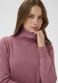 Ružičasta turtleneck pulover z ribastim ovratnikom in manšetami, izdelan iz mehkega materiala. Ima sproščen kroj z minimalno teksturo.