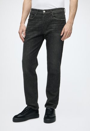 Homme portant un jean noir à jambes droites avec fermeture à boutons et des baskets noires, debout devant un fond blanc uni.