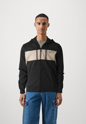 Felpa con zip nera e beige con cappuccio, caratterizzata da un pannello orizzontale centrale, tasche frontali e un piccolo logo verde sul petto.
