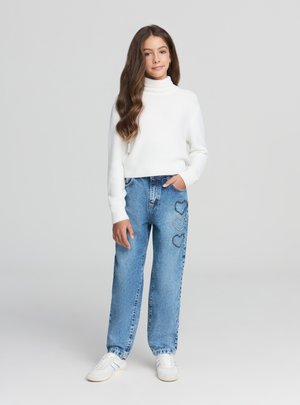 Dziewczyna w białym swetrze z golfem, niebieskich dżinsach z trzema wzorami serc na lewym udzie oraz białych trampkach, stojąca na jasnym tle.