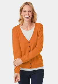 Oranje cardigan met een V-hals, lange mouwen en blauwe knopen, gedragen over een witte shirt; gemaakt van een zachte stof met een gladde textuur.