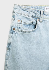 Gros plan sur un jean en denim bleu clair montrant la poche avant, la poche à monnaie, la passants de ceinture et les rivets en cuivre avec des détails de couture visibles.