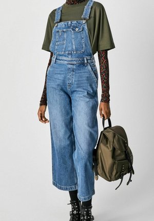 Osoba ubrana w niebieski denimowy kombinezon, oliwkowozieloną koszulę, długie kwieciste rękawy, czarne ćwiekowane buty, trzymająca oliwkowozielony plecak.